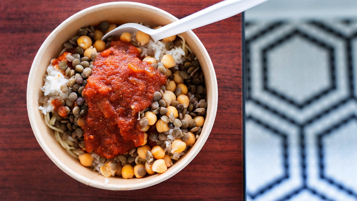 Un tazón de koshari, una comida vegetariana con capas de pasta, arroz, garbanzos, lentejas y salsa de tomate cubierta con cebollas fritas crujientes, vistas el jueves 10 de julio de 2025, se sirve con salsa de vinagre de ajo y salsa de chile caliente en Remo's Café, en Jackson, Miss. Se ve en mesa de madera con un piso azul y blanco en el fondo.