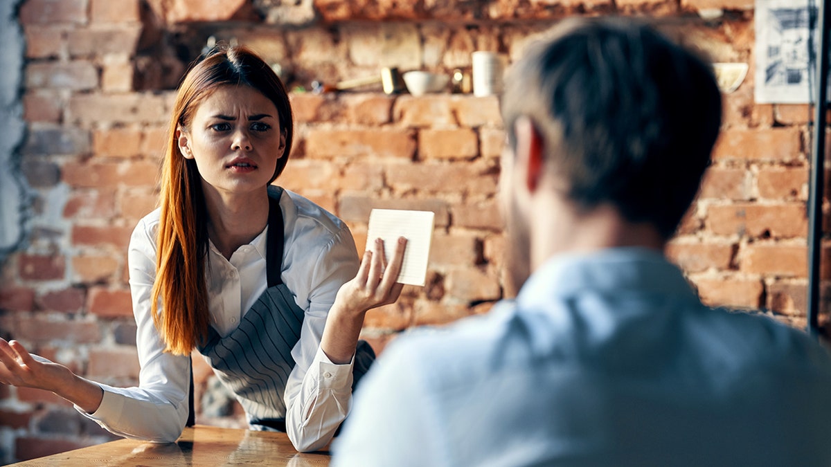A waitress at a restaurant holds up her hands and seems confused by a customer at a table.