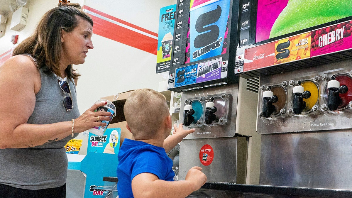 A woman and child stand in front of a Slurpee machine at a 7-Eleven.