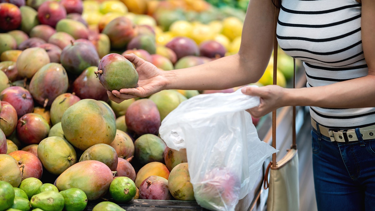 A woman holds a mango in a bag in one hand and another in her other hand.