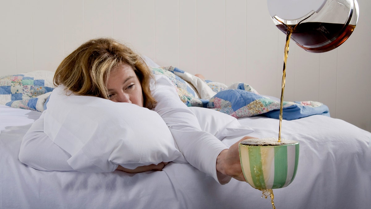 A woman in bed holds an overflowing coffee mug in her hand from the pot above.