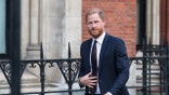 Prince Harry wearing a dark suit walking next to a fence with his hand on his stomach