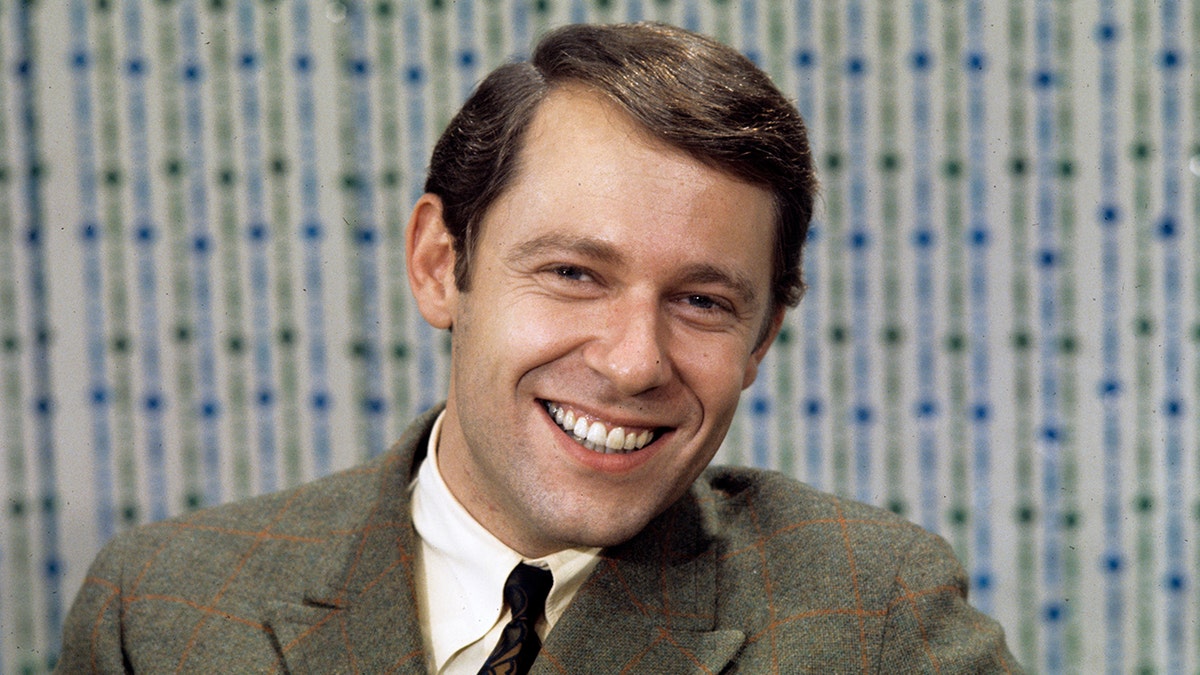A close-up of a young Alan Hamel smiling in a suit and tie.