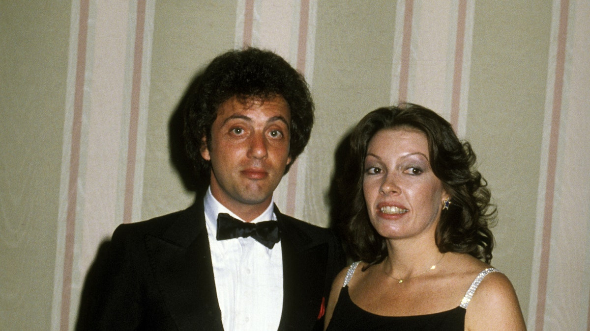 A young Billy Joel in a tux standing next to Elizabeth Weber wearing a sparkling spaghetti strap black dress.