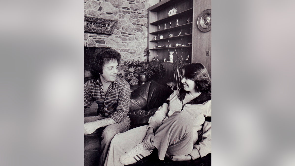 A black and white photo of Billy Joel and Elizabeth Weber smiling at each other inside a home.