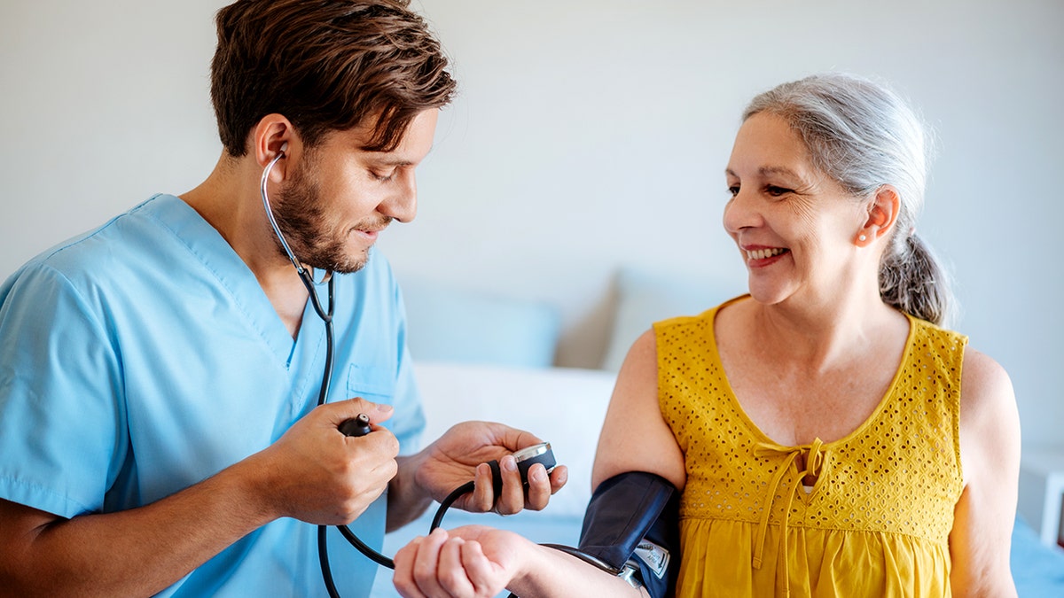 A nurse in the blue scrub checks blood pressure for a smiling woman.
