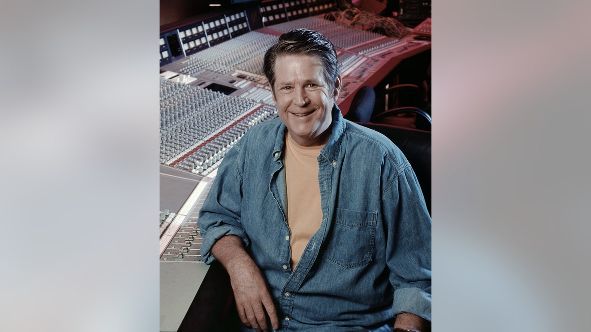 布萊恩·威爾遜（Brian Wilson）在工作室面前微笑著，穿著黃色T恤的牛仔布打開襯衫。