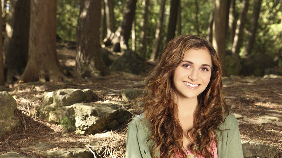 A close-up of Alyson Stoner smiling in a green jacket sitting in a forest.