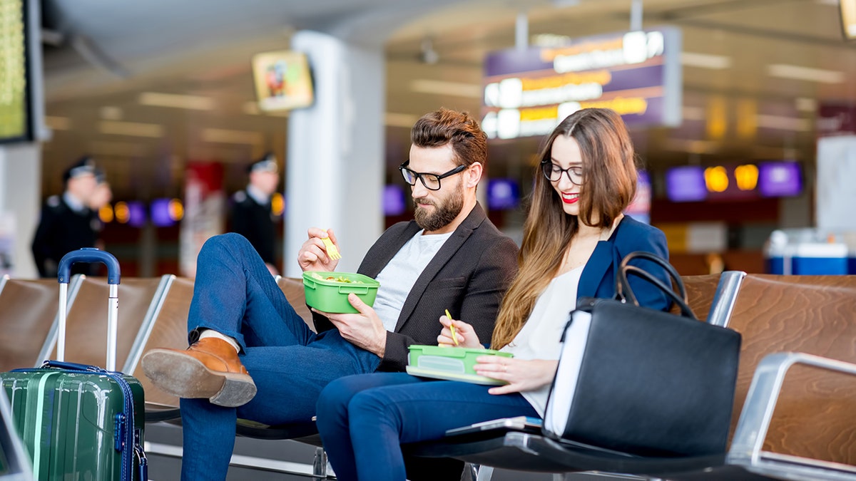 A man and a woman seated next to each other at an airport eat from bowls on their laps.