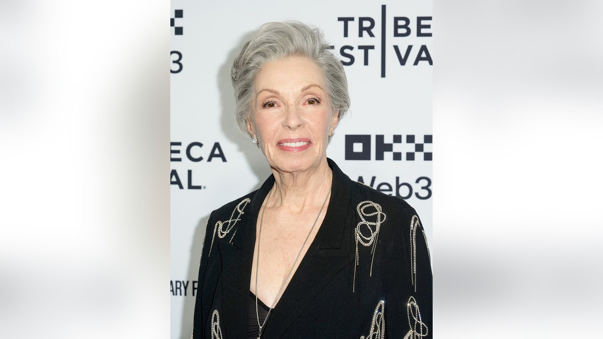 Elizabeth Weber wearing a low cut black suit dress with gold chains.