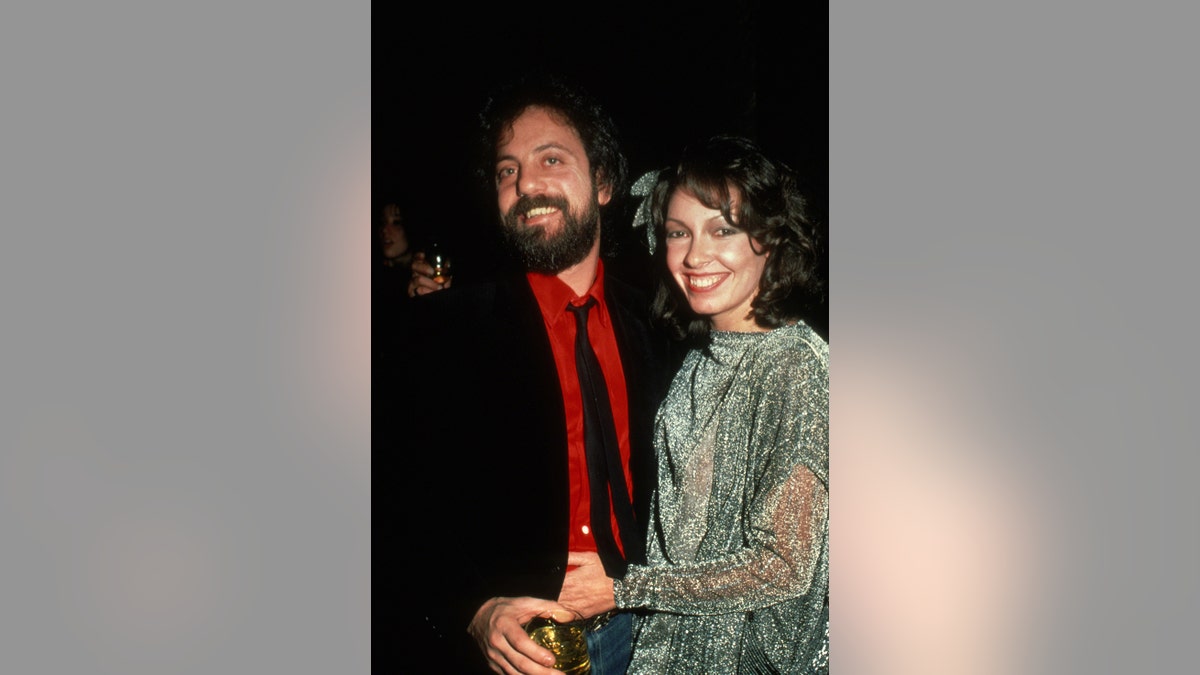 Billy Joel in a red shirt and a black blazer with a matching tie next to Elizabeth Weber smiling in a sea green sheer blouse.