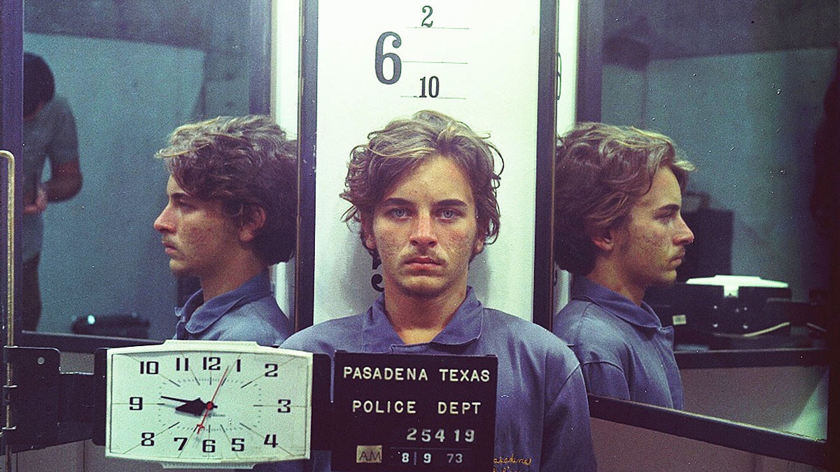 Elmer Wayne Henley wearing a blue shirt looking serious as he poses for his mugshot.