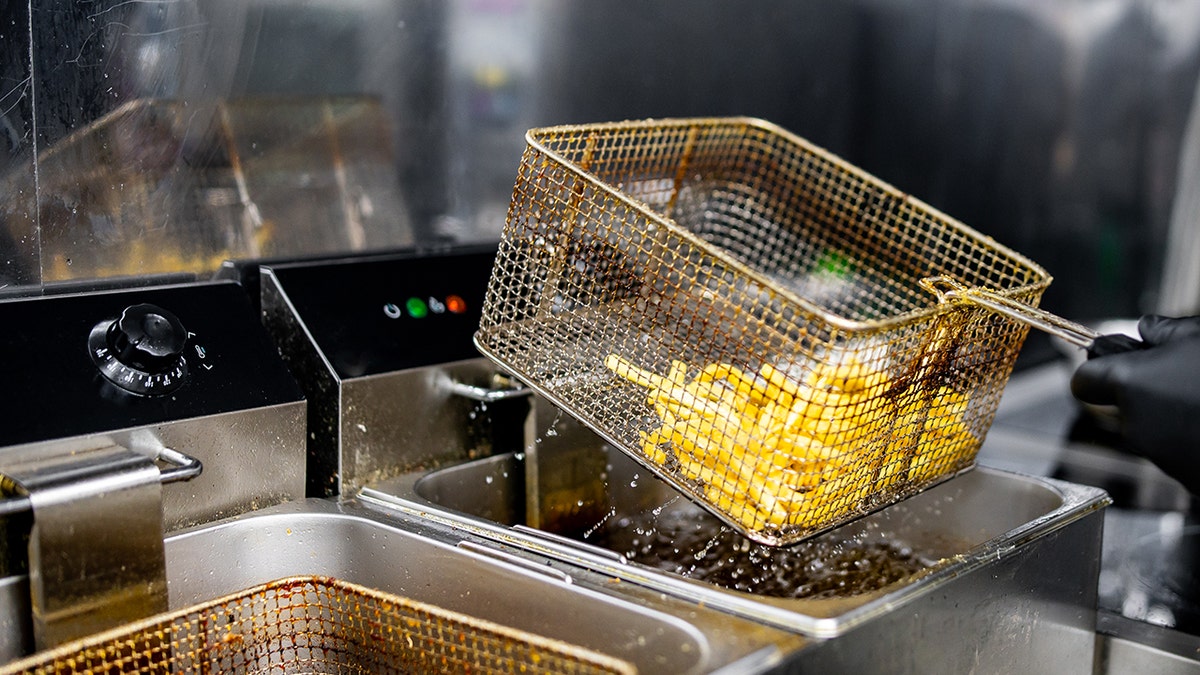 Worker with black gloves lifts a basket of freshly fried fries from a commercial deep fryer. Droplets of oil capture the motion.