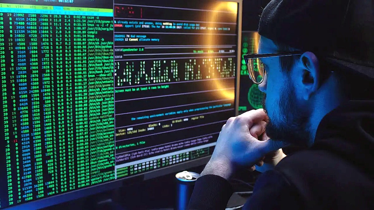 A man in the computer code stares on Hiis Monitor in a dark room, with a ring light that is reflected on the screen and canned drinks open next to it.