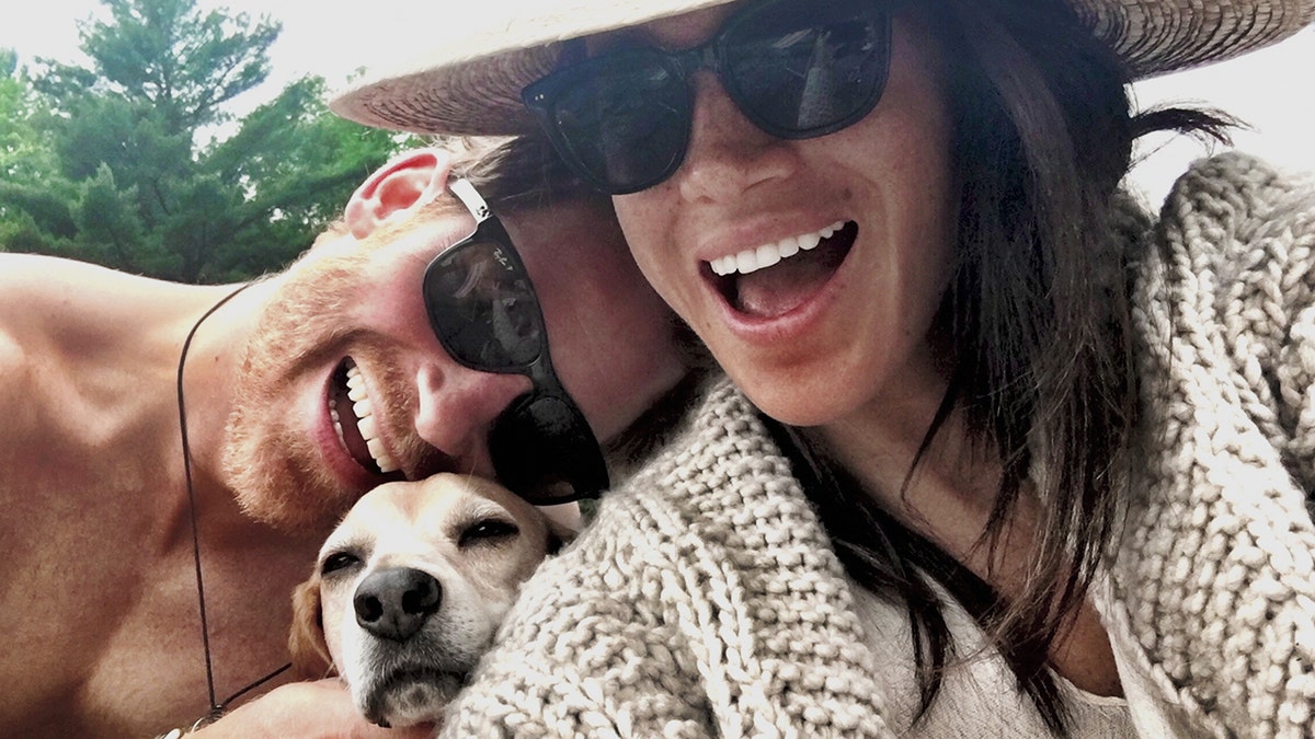 A close-up of Meghan Markle and Prince Harry wearing sunglasses and smiling while holding their dog.