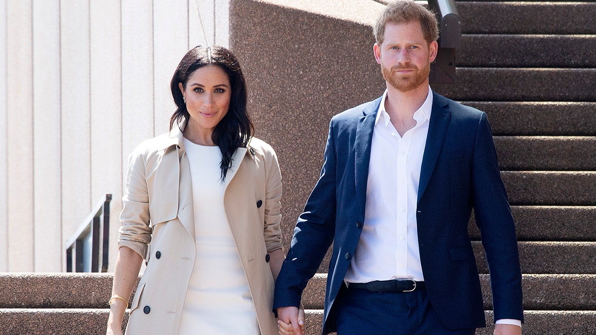 Prince Harry and Meghan Markle walking together looking serious and holding hands.