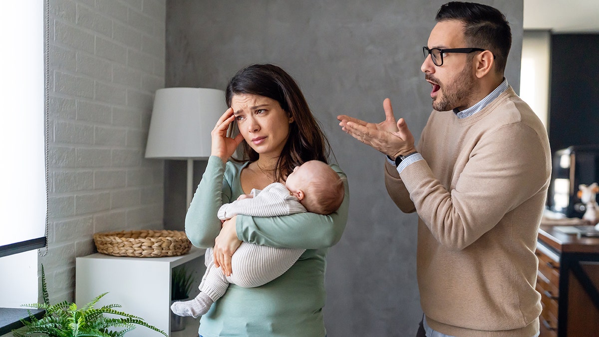 Husband arguing with wife as she holds baby and hand up to her head looking distressed