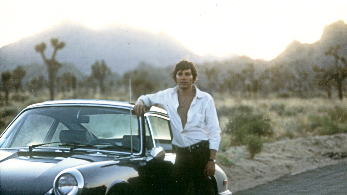 Jay Sebring wearing a white button down shirt in front of a car.