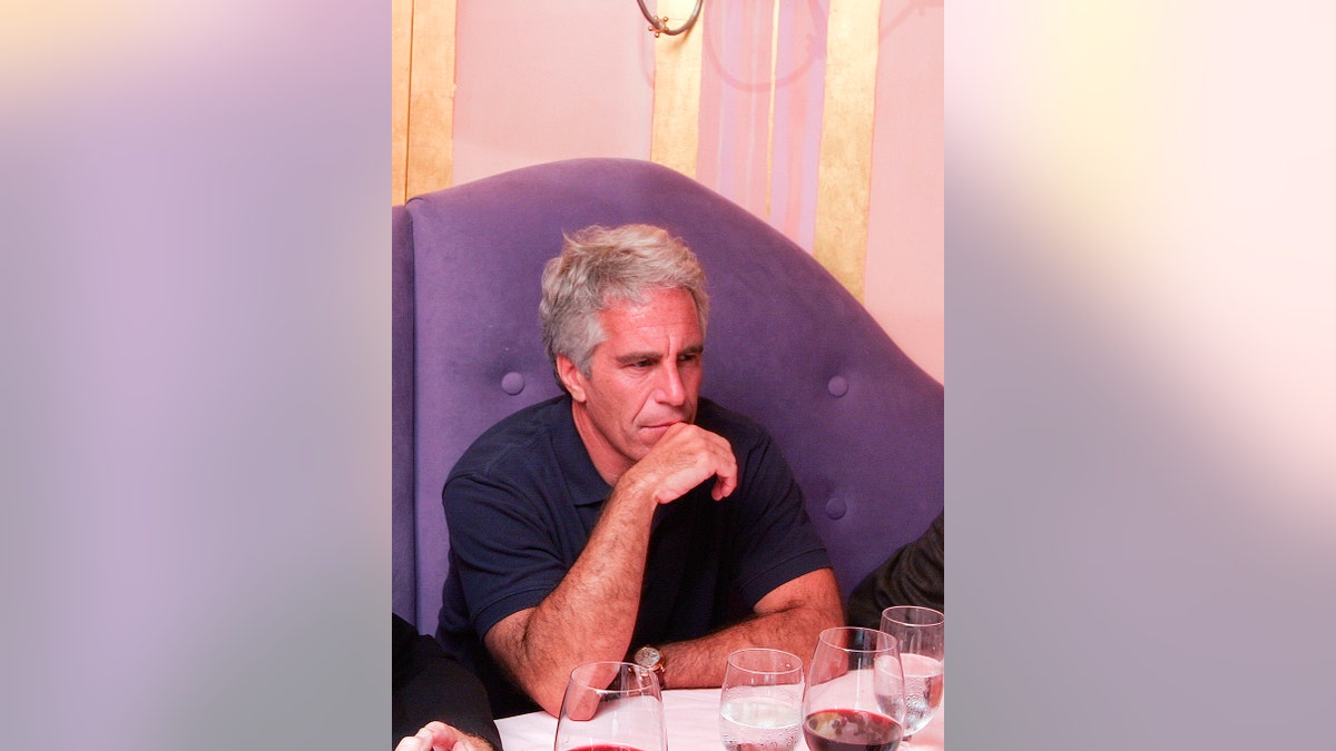 Jeffrey Epstein wearing a navy blue shirt sitting in a purple couch in front of a table.