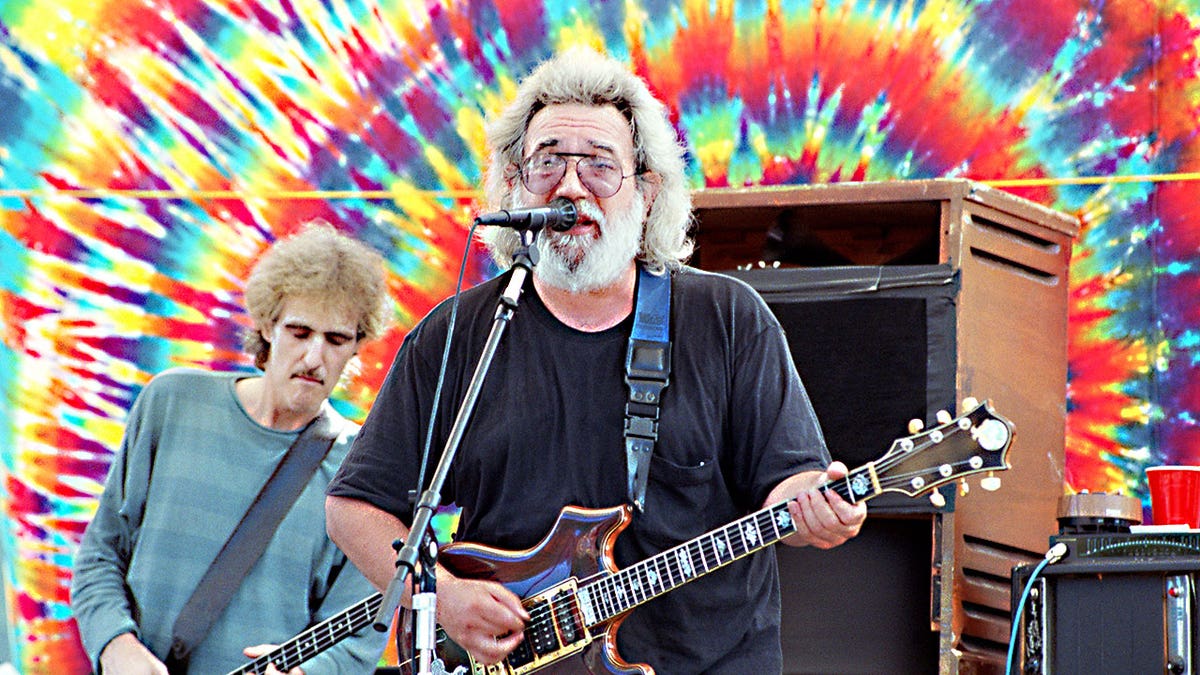 Jerry Garcia playing guitar with the Grateful Dead on stage against a bright tie dye background.