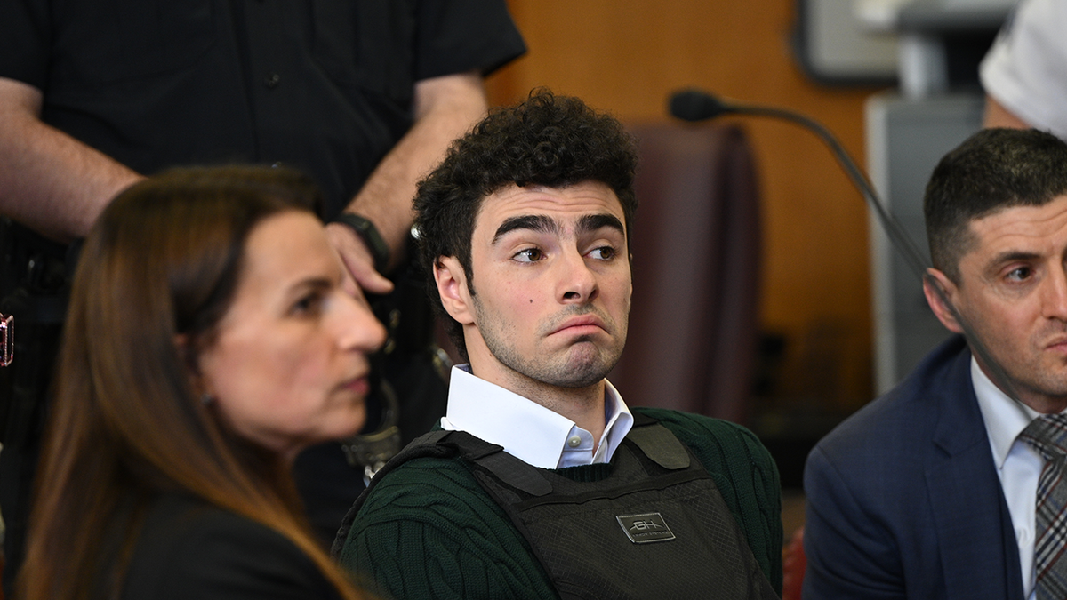 A curly haired Luigi Mangione, wearing a bulletproof vest over his green sweater and collared shirt, makes a facial expression during a hearing in Manhattan.
