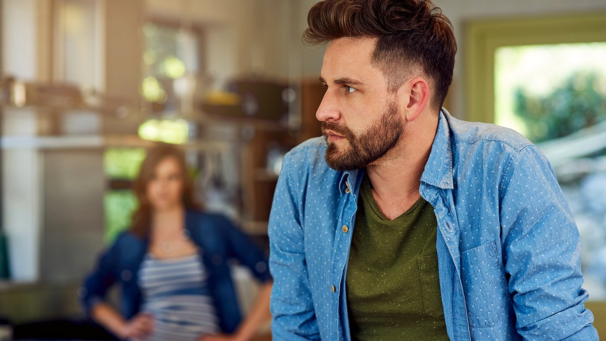A man is annoyed with a woman who is out of focus in a kitchen.