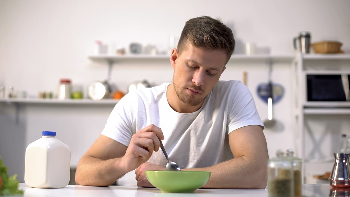 A man wearing a white T-shirt looks down at his green cereal bowl while holding a silver spoon.
