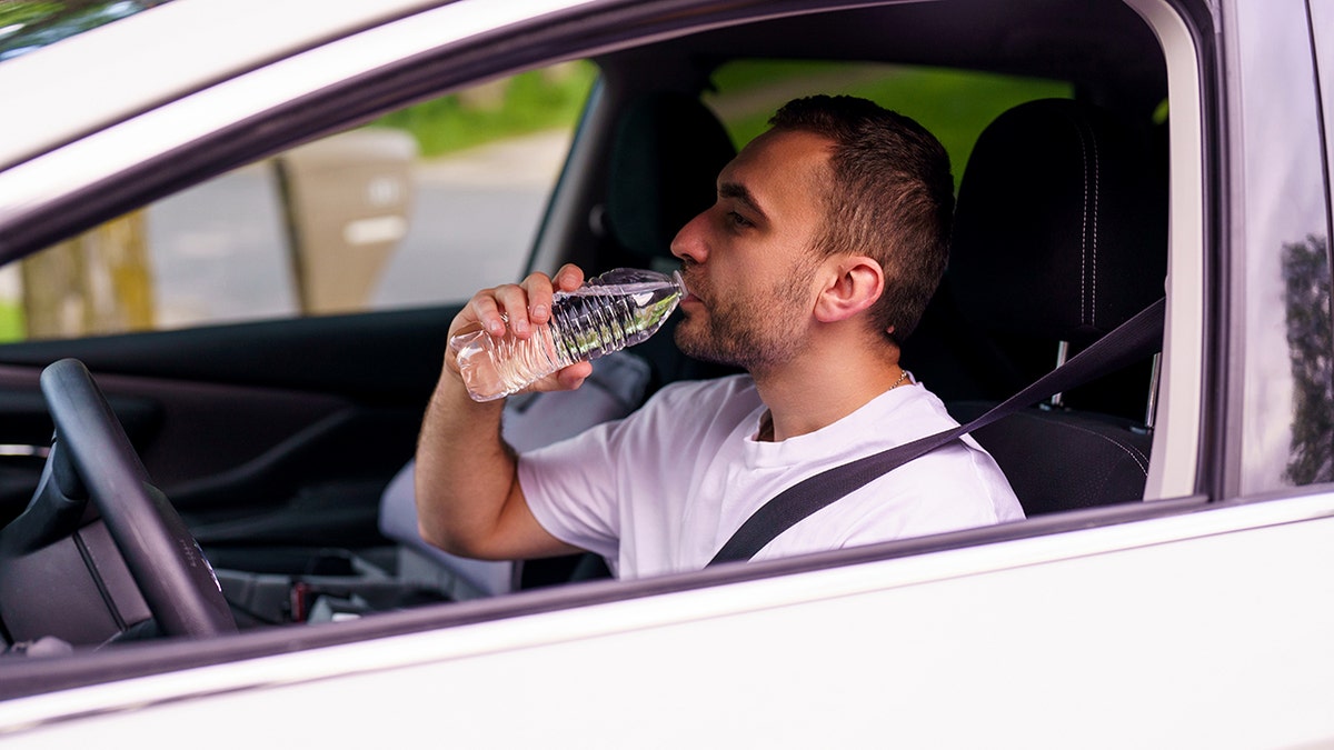 Vīrietis dzer no plastmasas ūdens pudeles baltas automašīnas vadītāja sēdeklī.