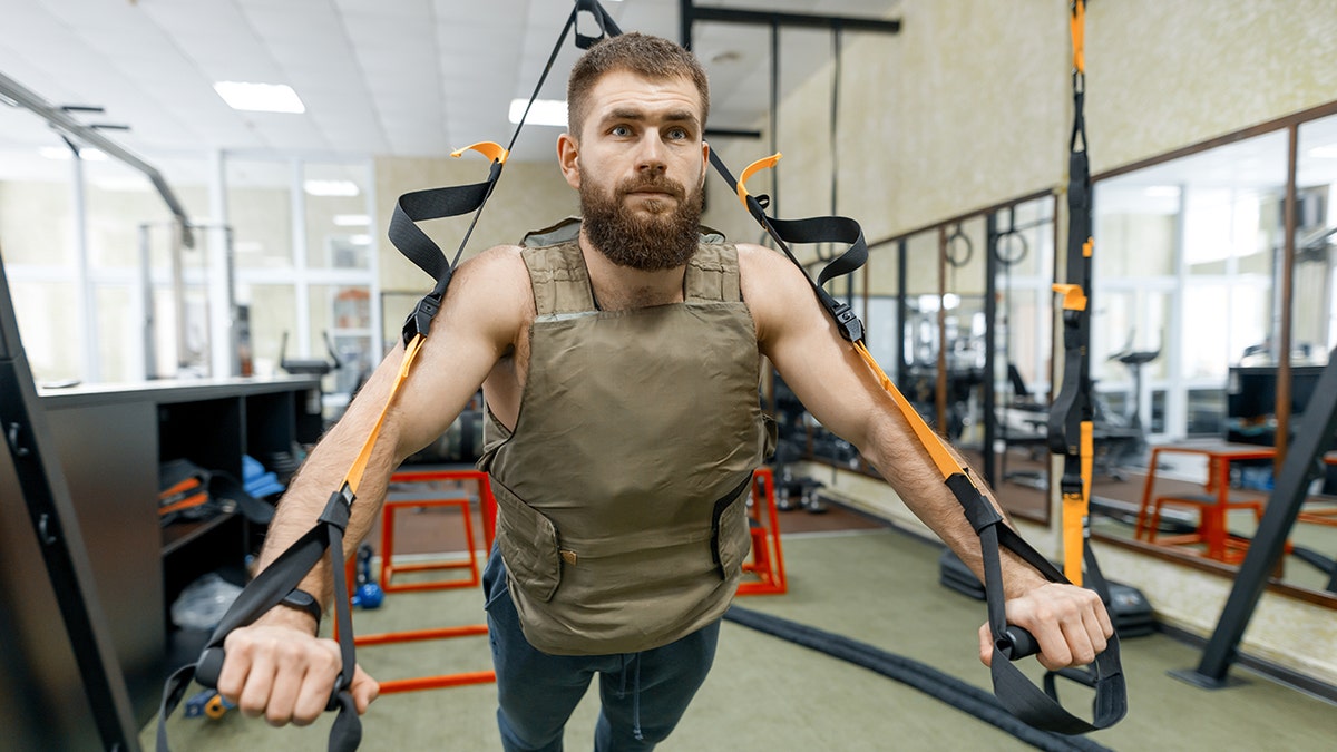 A man exercises out in gym using TRX cables and wearing weighted vest, a new fitness trend to help boost workouts.
