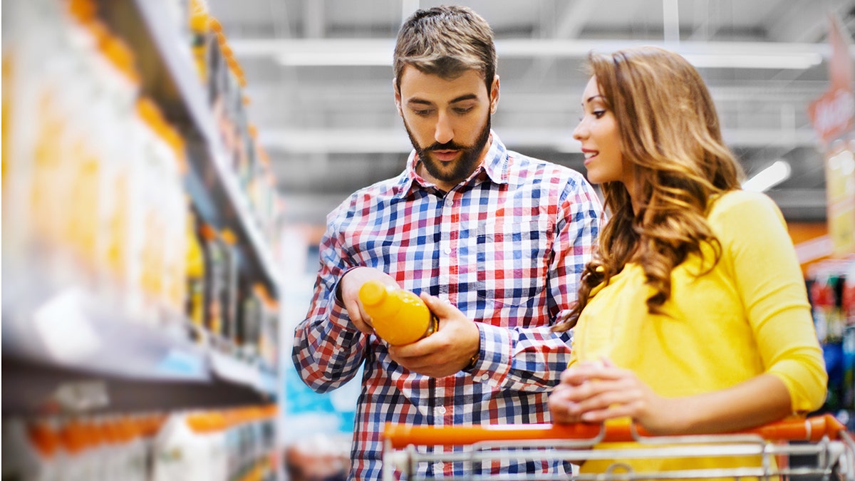 Man and woman looking at orange juice label at grocery store, shopping cart in front of them and juices on shelves in background