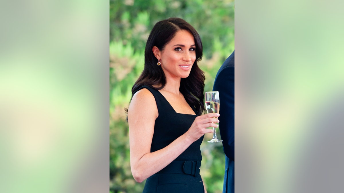 Meghan Markle wearing a strappy black dress holding a champagne flute.