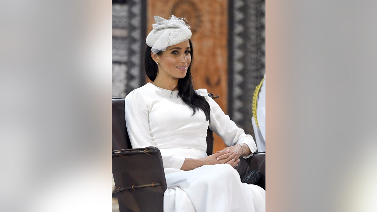 Meghan Markle valkā baltu kleitu ar atbilstošu fascinatoru smaidot.