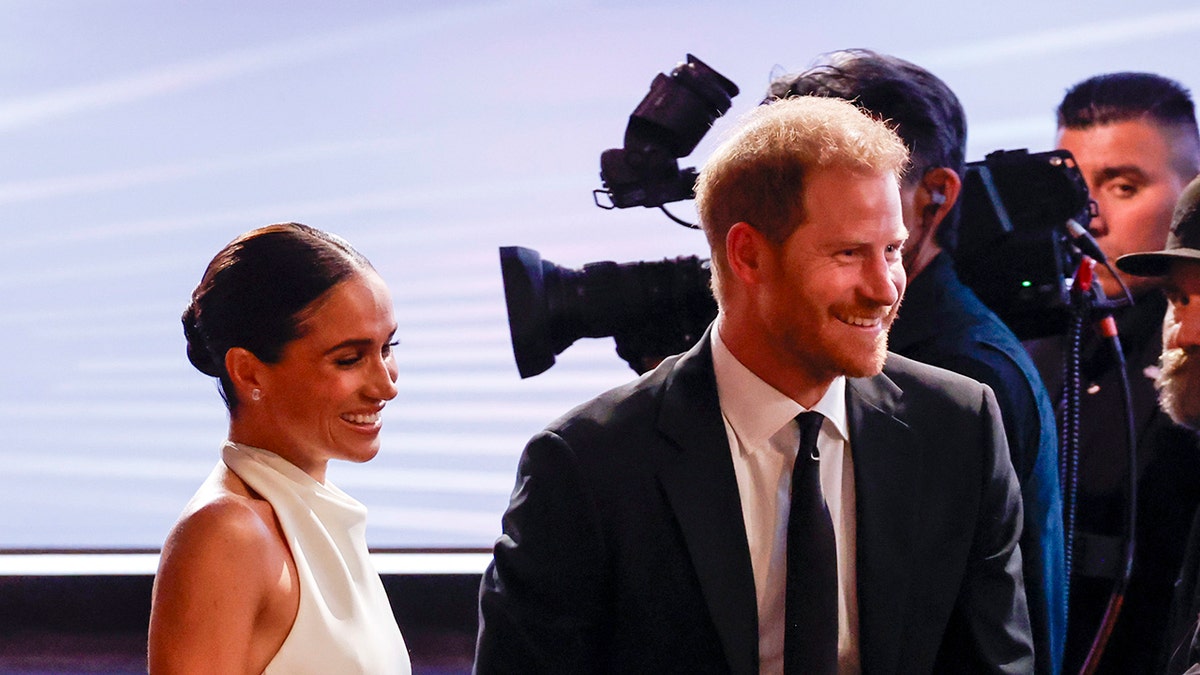 Meghan Markle wearing a strapless white dress walking behind Prince Harry in a suit with cameras behind them.