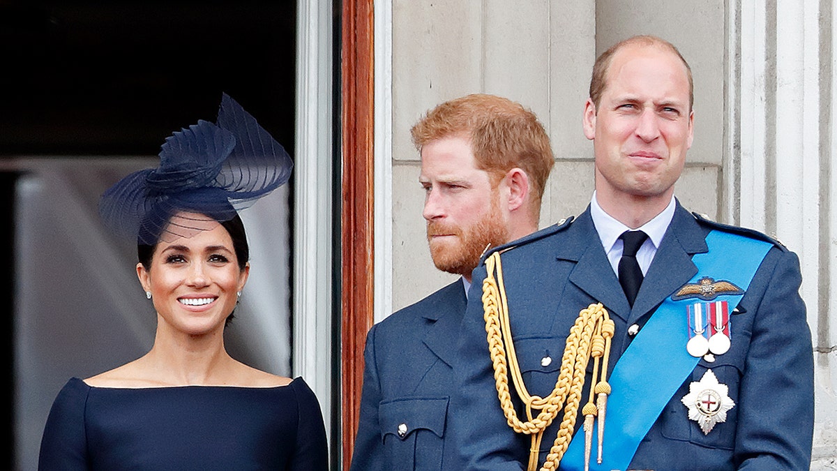 Meghan Markle smiling in a blue dress with a matching fascinator with Prince Harry looking ahead on and Prince William in a suit with medals standing in front of them.