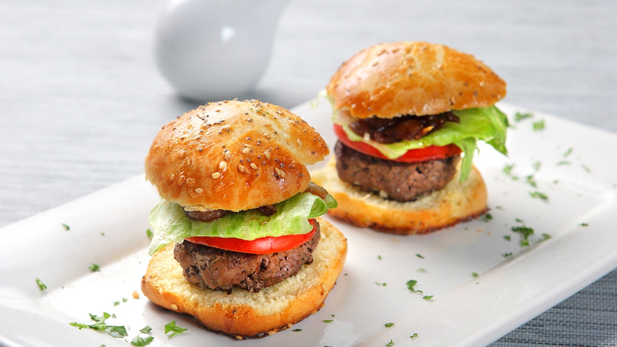 Two burger sliders seen on plate, garnished with tomatoes, lettuce, bacon and parsley