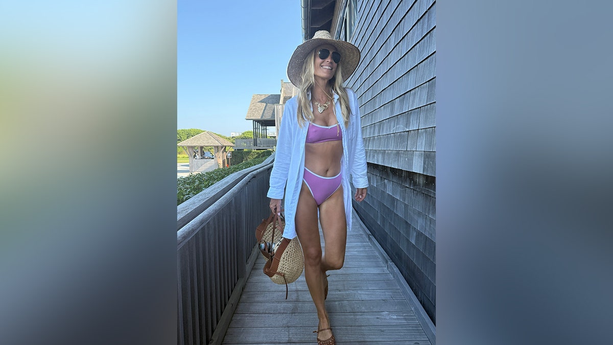 Molly Sims smiling at the camera while dressed in a purple bikini and white shirt.