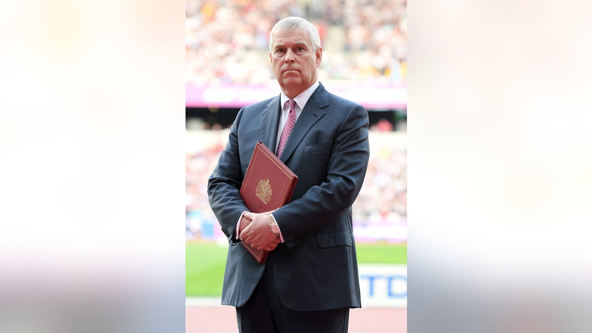 Prince Andrew wearing a dark suit and red tie holding a burgundy book.
