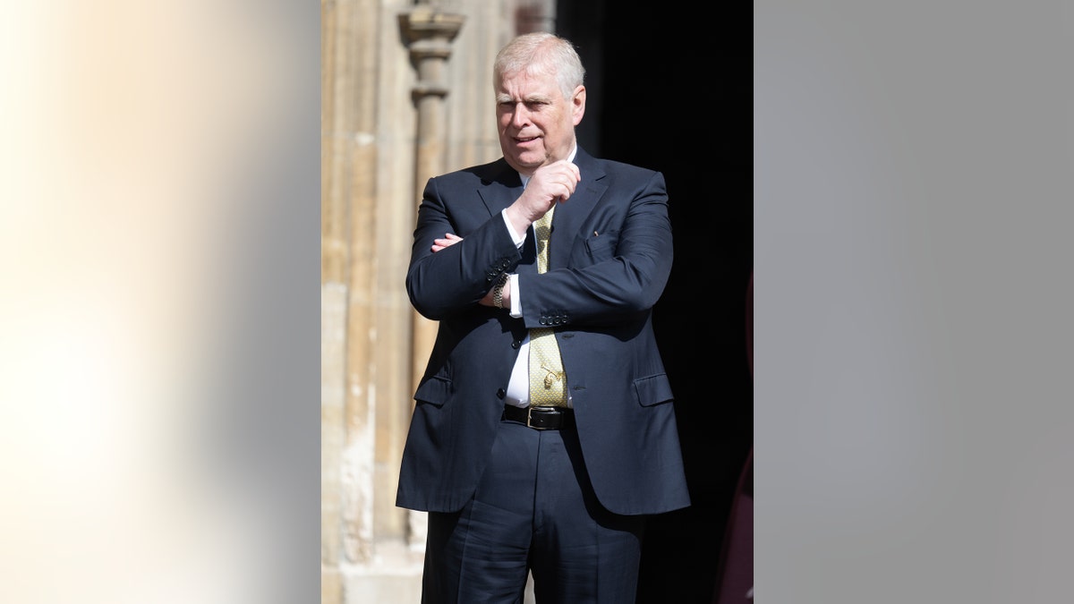 Prince Andrew smirking in a dark suit with a yellow tie while standing outdoors.