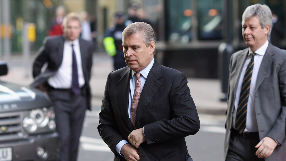 Prince Andrew wearing a dark suit walking outside ahead of two men watching.