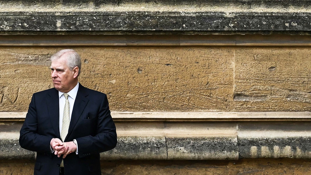 Prince Andrew wearing a dark suit looking serious in front of a stone wall.