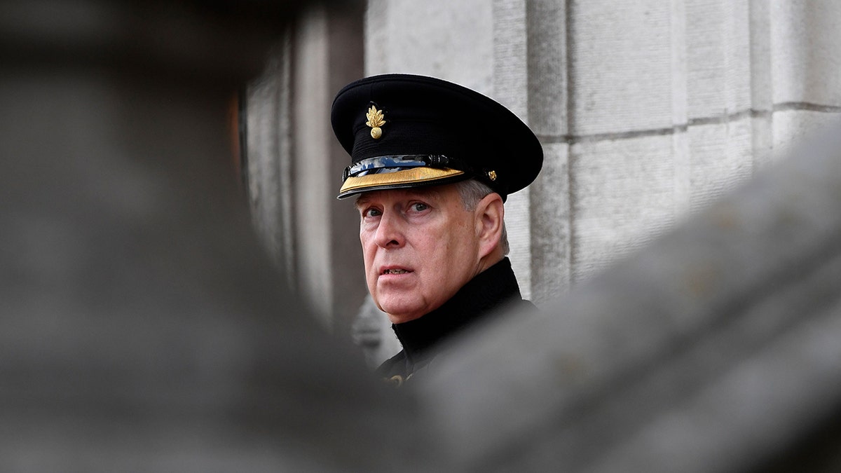 Prince Andrew wearing a military uniform and hat looking downcast behind a grey building.