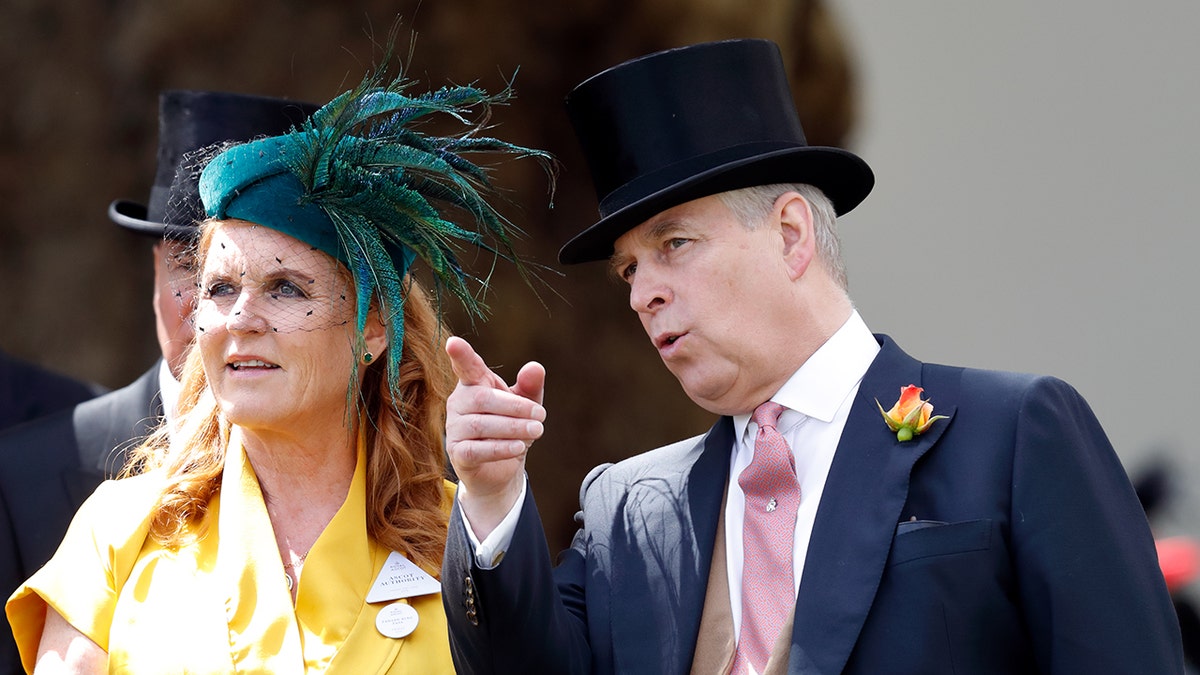 Prince Andrew in a dark blue suit and black top hat point up as Sarah Ferguson, wearing a yellow dress and green fascinator, looks on.