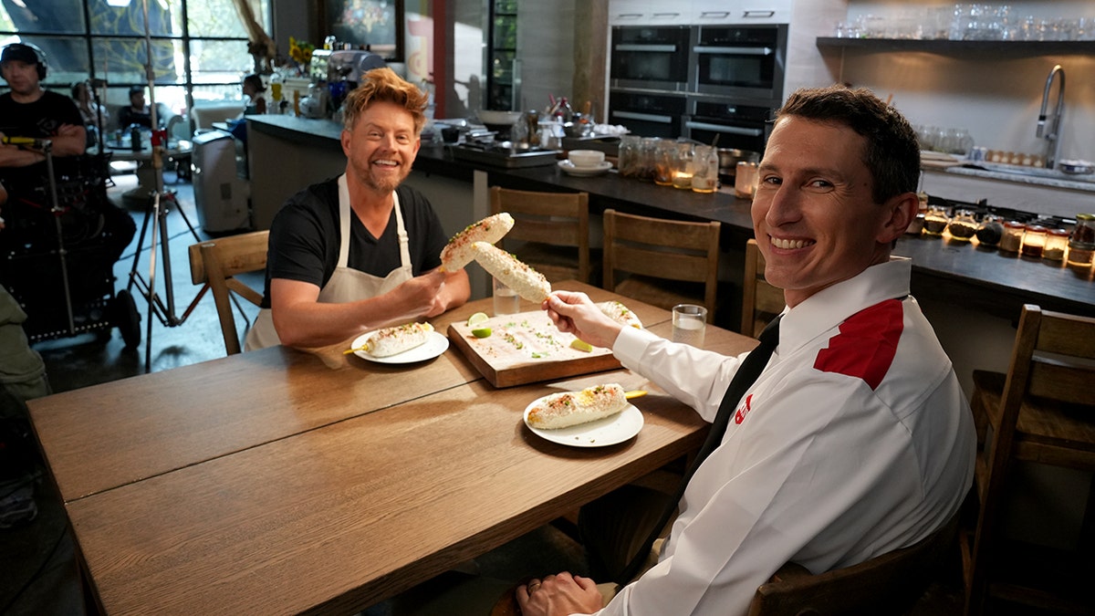 Richard Blais and a man wearing an Orkin uniform hold food in their hands at a table.