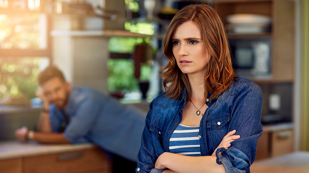 A woman stares away from the camera, looking annoyed as a man, who is out of focus, slumps over a counter in a kitchen.