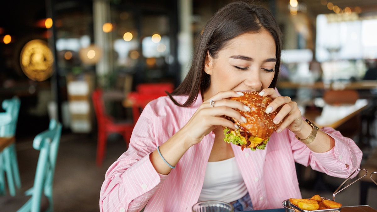 Young woman dining in fast food restaurant, biting into burger with juice and sides in front of her.
