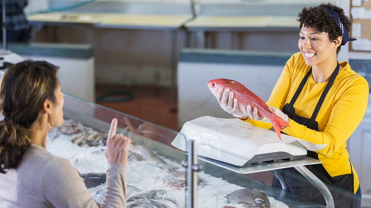 Women in seafood market helping clients, women shopping problem seen from behind while workers pull out big pink fish