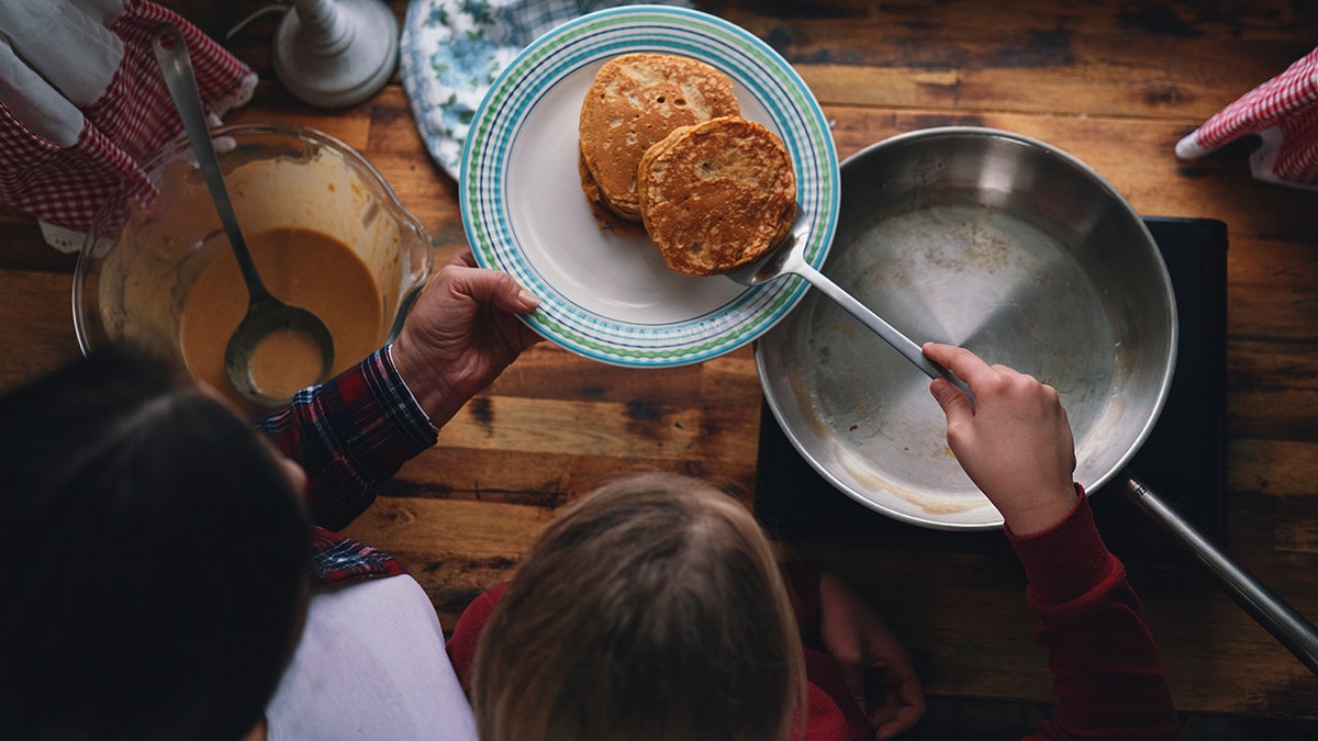 Mother and Daughter making Pancakes, seen from above in country-style kitchen; transferring pancakes from pan to stack on plate
