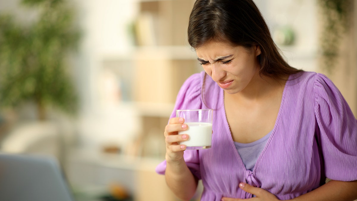 A woman holds her stomach and appears in pain while holding a glass of milk.