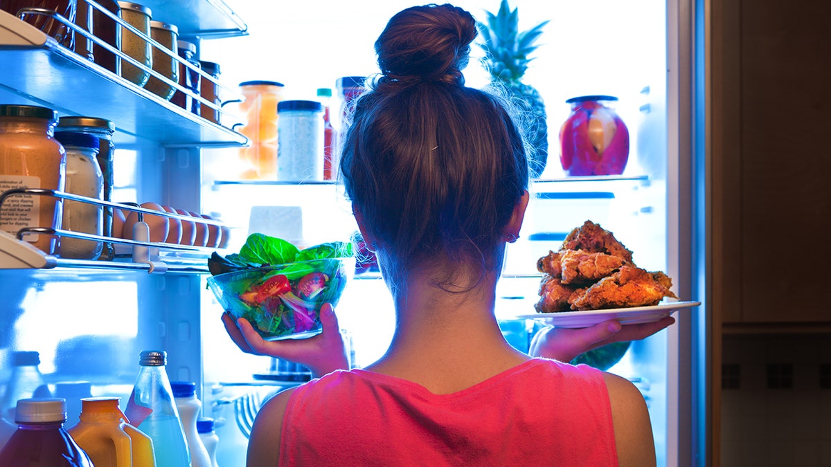 Woman seen from behind standing in front of fridge holding salad and fried chicken, indicating choosing healthy or junk food