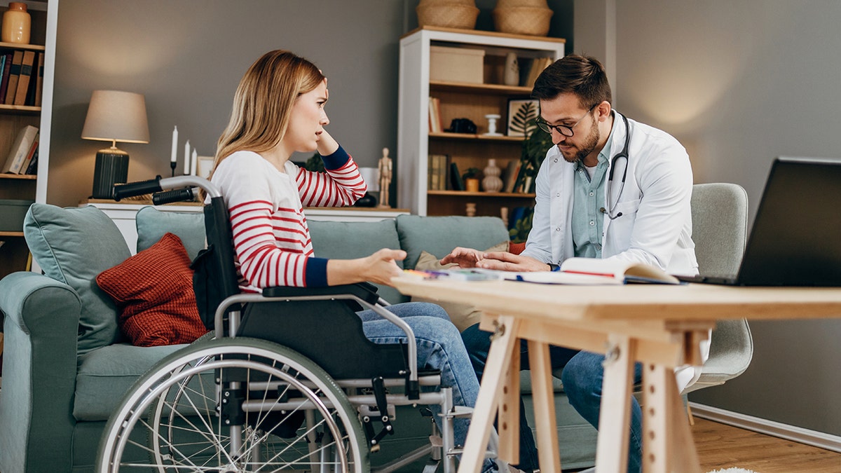 Woman seen in wheelchair during check-up with doctor. She holds her head as doctor appears to explain something to her